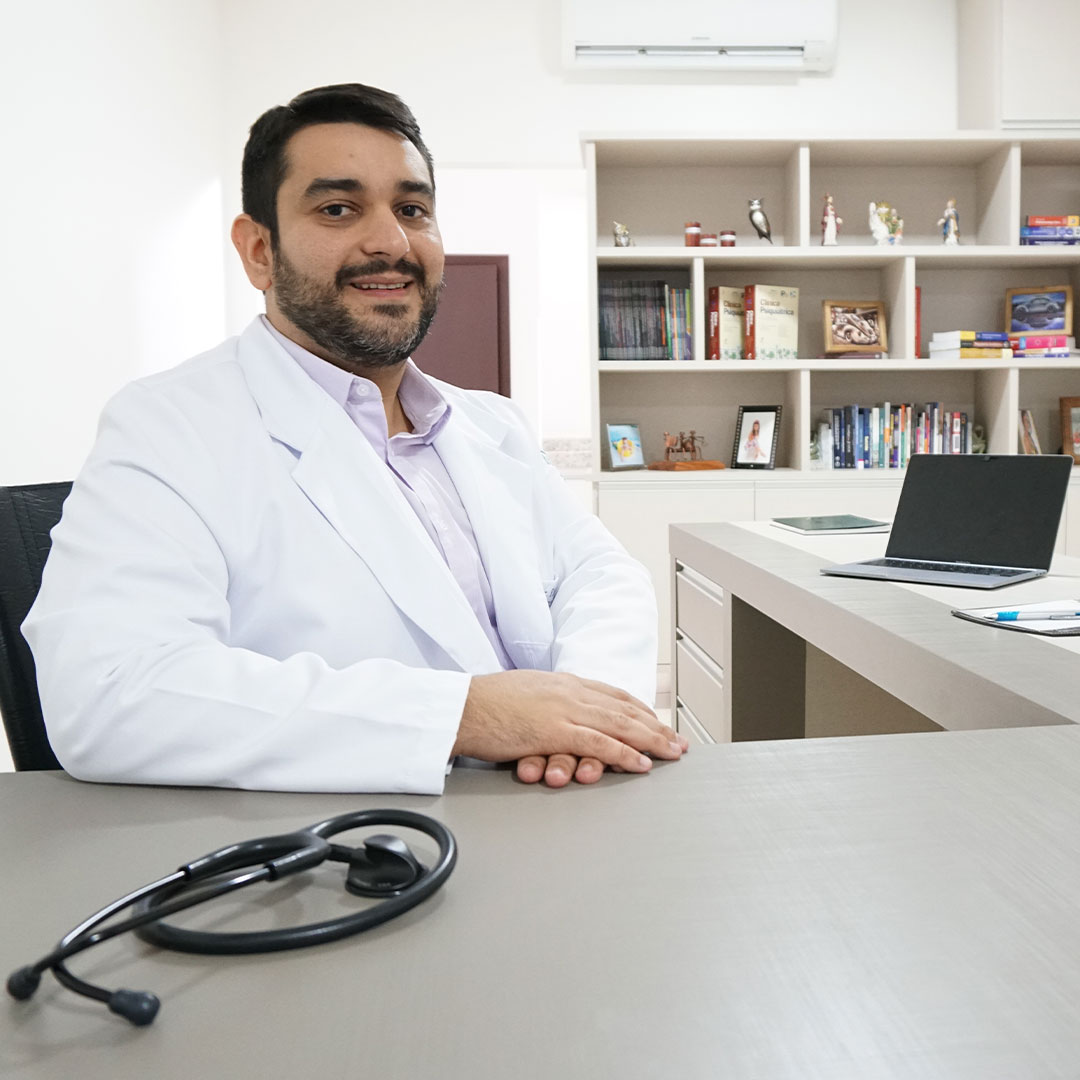 Dr. Rafael Gonçalves Lima, psiquiatra, sentado à mesa do consultório em Corinto, com a estante de livros de psiquiatria ao fundo.